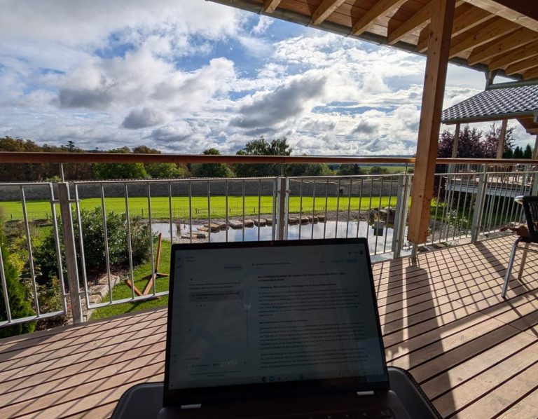 Blick auf eine grüne Wiese und den Himmel von einer Terrasse mit Laptop im Vordergrund.