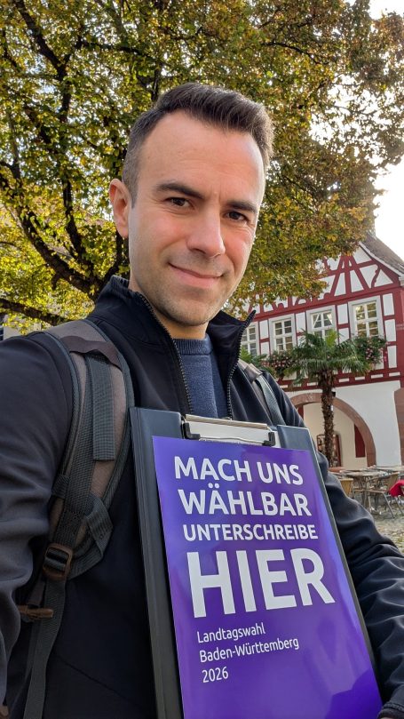 Mann mit einem Schild, auf dem "Mach uns wählbar" steht, vor historischer Gebäudefassade.